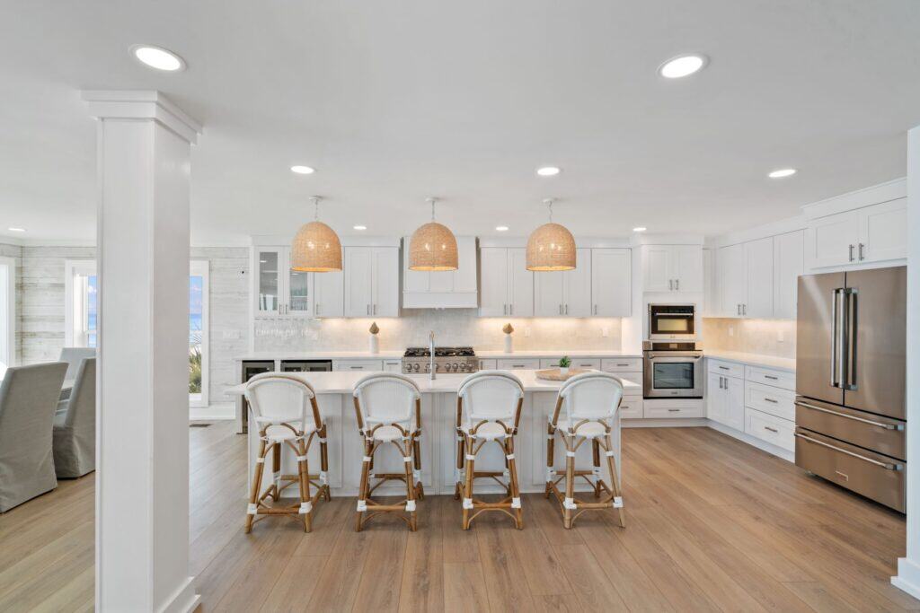 A spacious and modern kitchen features white cabinetry, wooden flooring, pendant lights, and wicker barstools at a central island, with a dining area nearby.