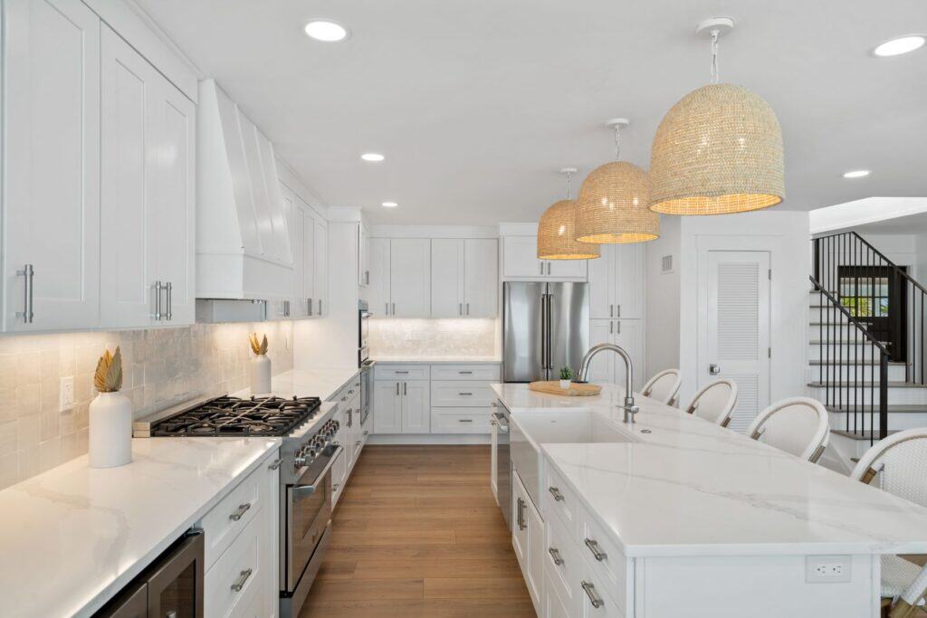 Spacious modern kitchen with white cabinetry, marble countertops, stainless steel appliances, wicker pendant lights, and wooden flooring, adjacent to a staircase.