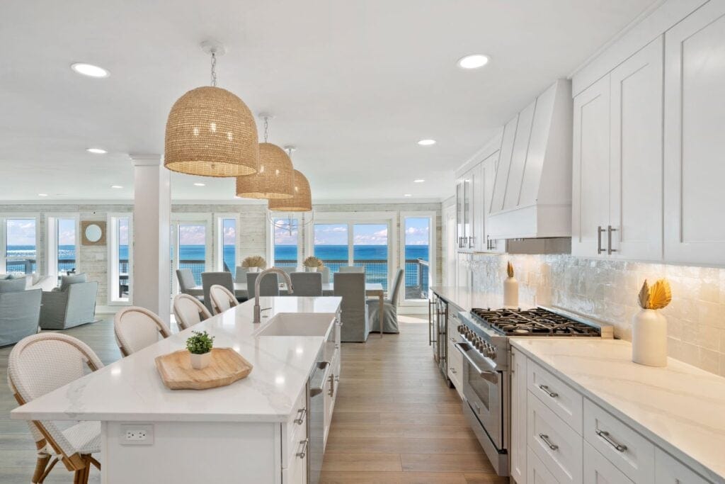 Modern coastal kitchen with white cabinets, large island, wicker pendant lights, and ocean view through wide windows, featuring a dining area and lounge.
