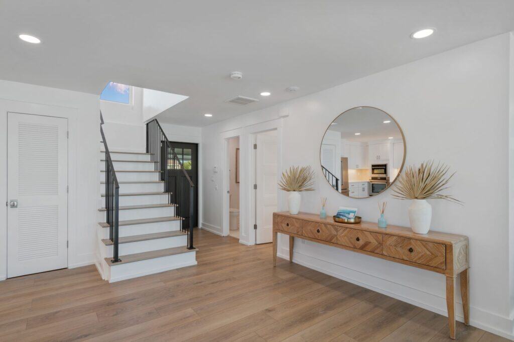Bright, modern interior with wooden floors, white walls, and a staircase. A console table with décor is below a round mirror.