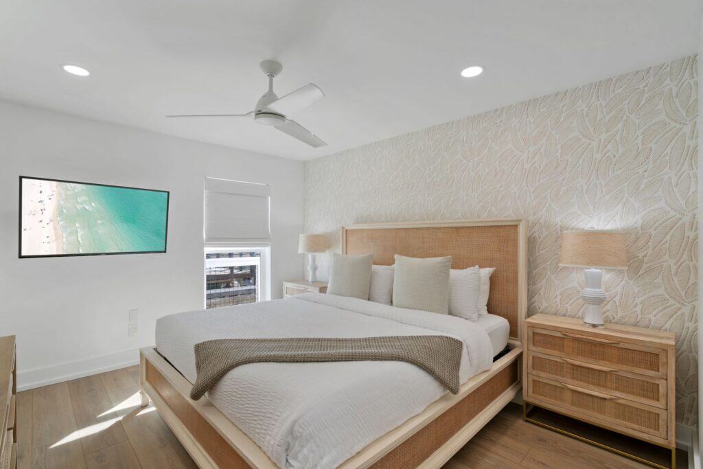 A modern bedroom with white walls, stylish wallpaper, a king-sized bed, wooden furniture, and a TV displaying a beach scene.