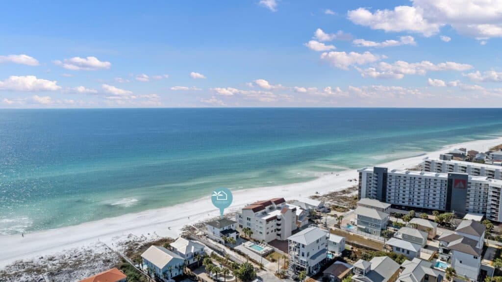 Aerial view of a picturesque coastline with turquoise waters, sandy beach, and modern buildings. Clear blue sky with scattered clouds overhead.