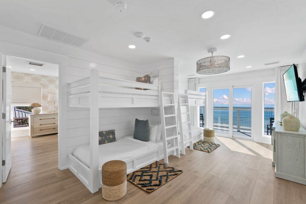 Bright, modern room with bunk beds, wooden flooring, and ocean view. Sliding glass doors lead to balcony; adjacent room visible through open doorway.