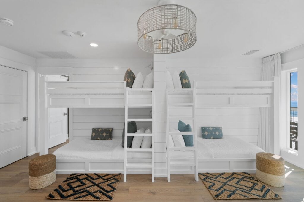 Bright room with white bunk beds, wooden floor, patterned rugs, woven stools. Large window shows ocean view, enhancing the airy, coastal decor.
