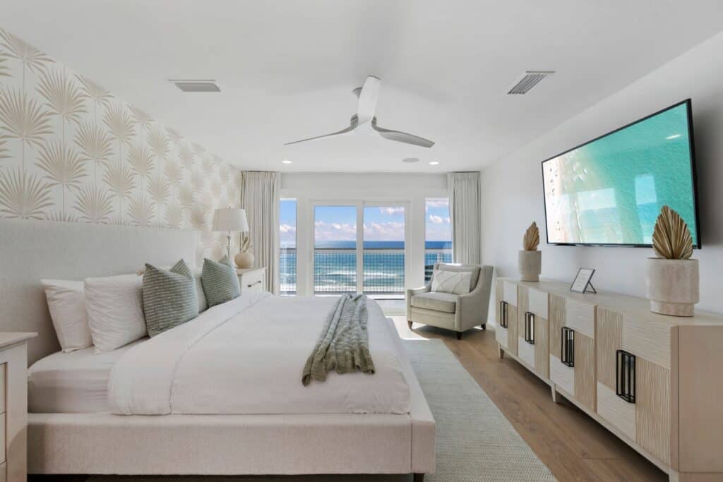 Modern bedroom with ocean view, featuring a large bed, patterned wallpaper, ceiling fan, and balcony doors. A serene, welcoming atmosphere.