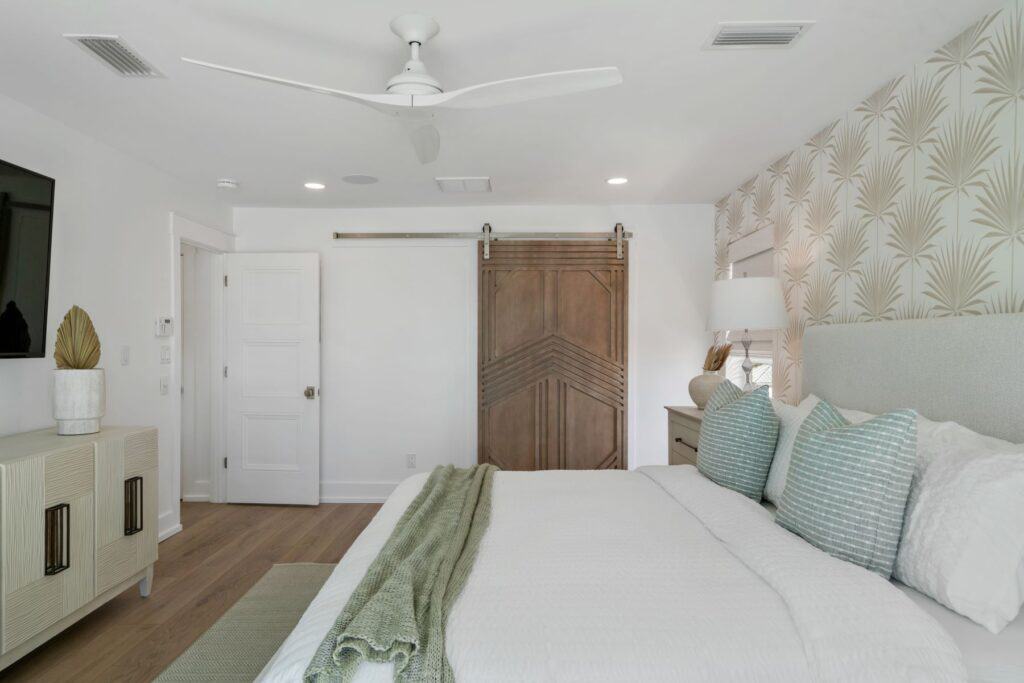Modern bedroom with a ceiling fan, patterned wallpaper, sliding barn door, and decorative throw on a neatly made bed.