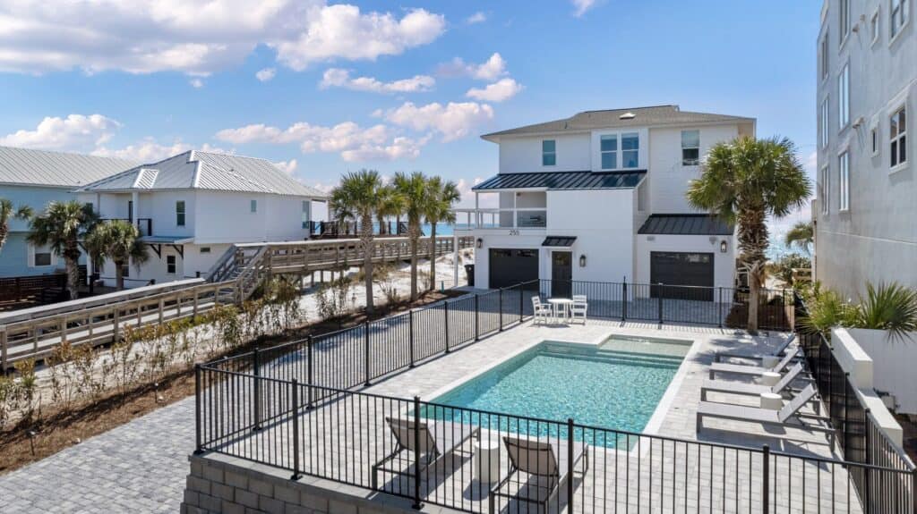Coastal scene with modern beachfront houses, palm trees, private pool, lounge chairs, and ocean view under a blue sky with scattered clouds.