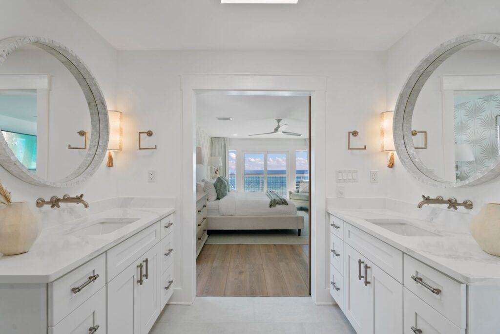 Modern bathroom with dual sinks and circular mirrors, leading to a bright bedroom with ocean views through large glass doors.