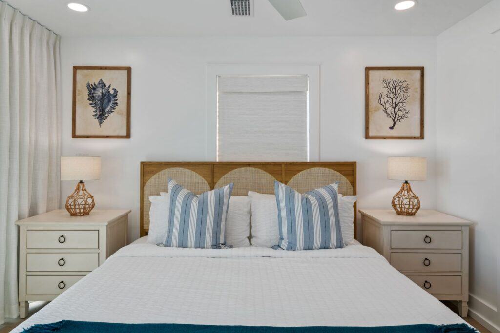 A cozy bedroom with a neatly made bed, striped pillows, bedside tables, wicker lamps, and coastal-themed artwork on white walls.