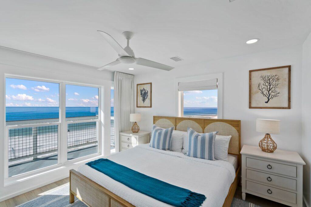 Bright bedroom with ocean view, featuring a king-sized bed, elegant decor, coastal-themed artwork, and large windows overlooking a serene beach.