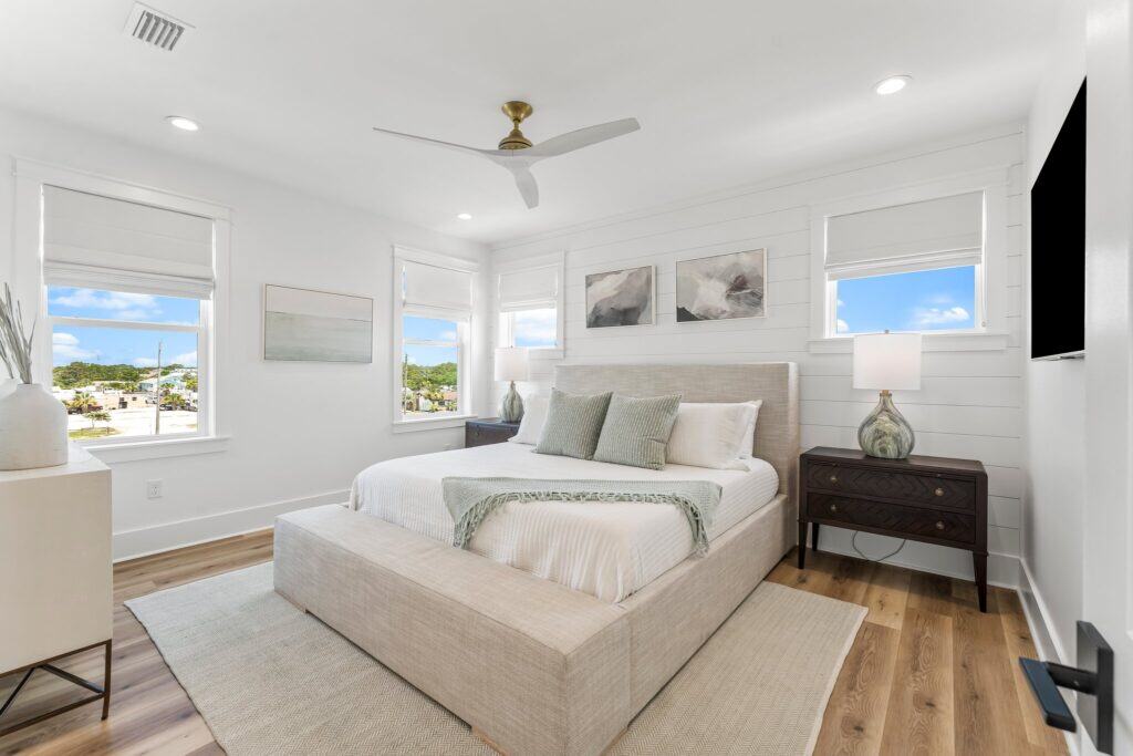 Bright bedroom with neutral colors, modern decor, and large windows showing outdoor greenery. Contains bed, nightstands, lamps, and abstract art. Ceiling fan above.