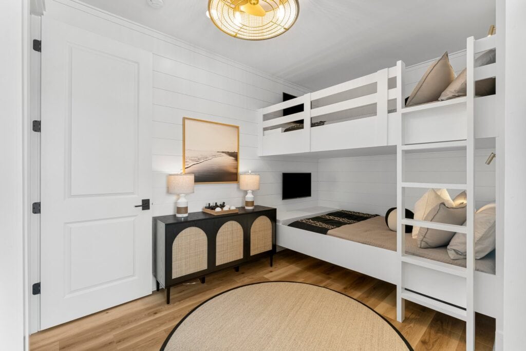 Cozy bedroom with white bunk beds, modern lighting, and a black cabinet. Minimalist decor features a beach photo and two lamps.