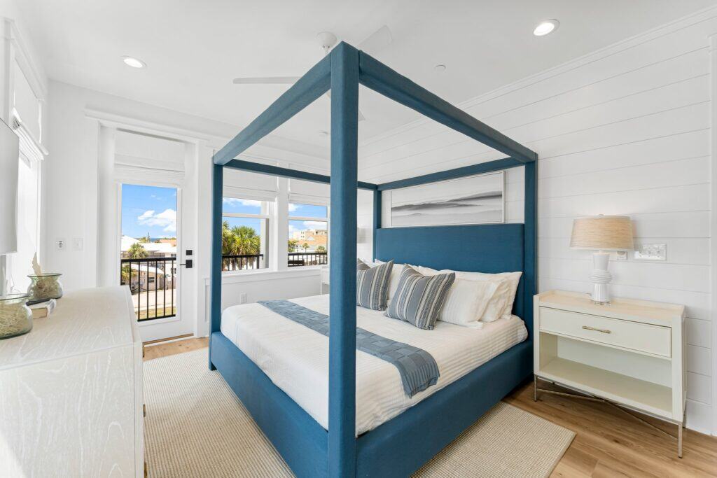 Bright bedroom with a blue four-poster bed, striped pillows, white walls, and a sunny view. Modern decor enhances the cozy atmosphere.