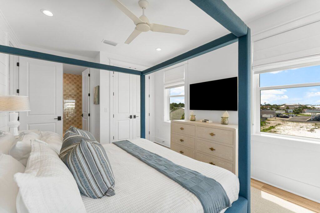 Modern bedroom with a canopy bed, ceiling fan, and flat-screen TV. Large window reveals an outdoor view of a suburban neighborhood.
