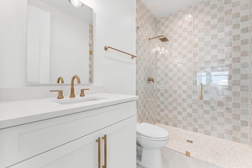 Modern bathroom features white vanity, brass fixtures, large mirror, and tiled walk-in shower with glass door, creating a clean, elegant space.