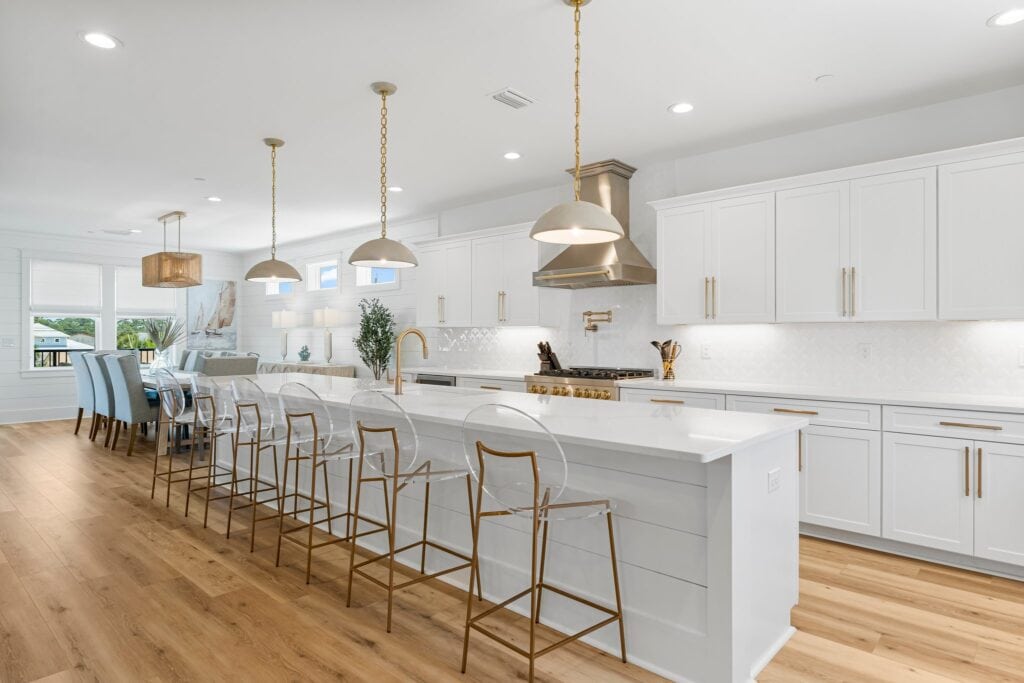 Spacious modern kitchen with wooden floors, white cabinets, large island, and transparent bar stools. Dining area features a long table and chairs.