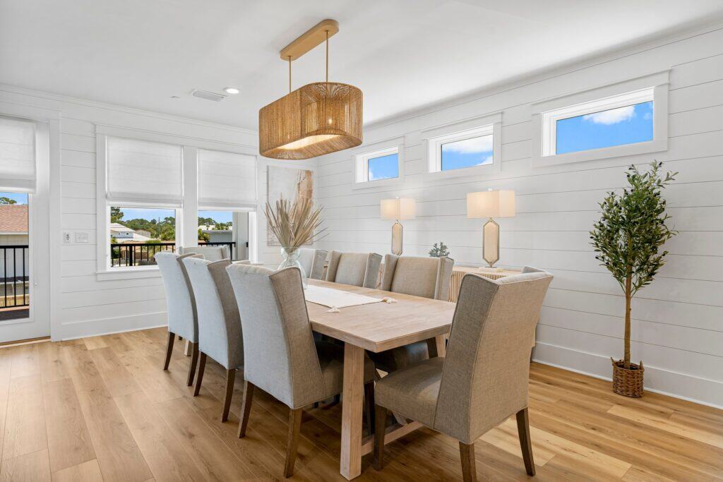 Bright dining room with wooden table, cushioned chairs, and modern pendant light. Large windows offer outdoor view; potted plants add greenery.