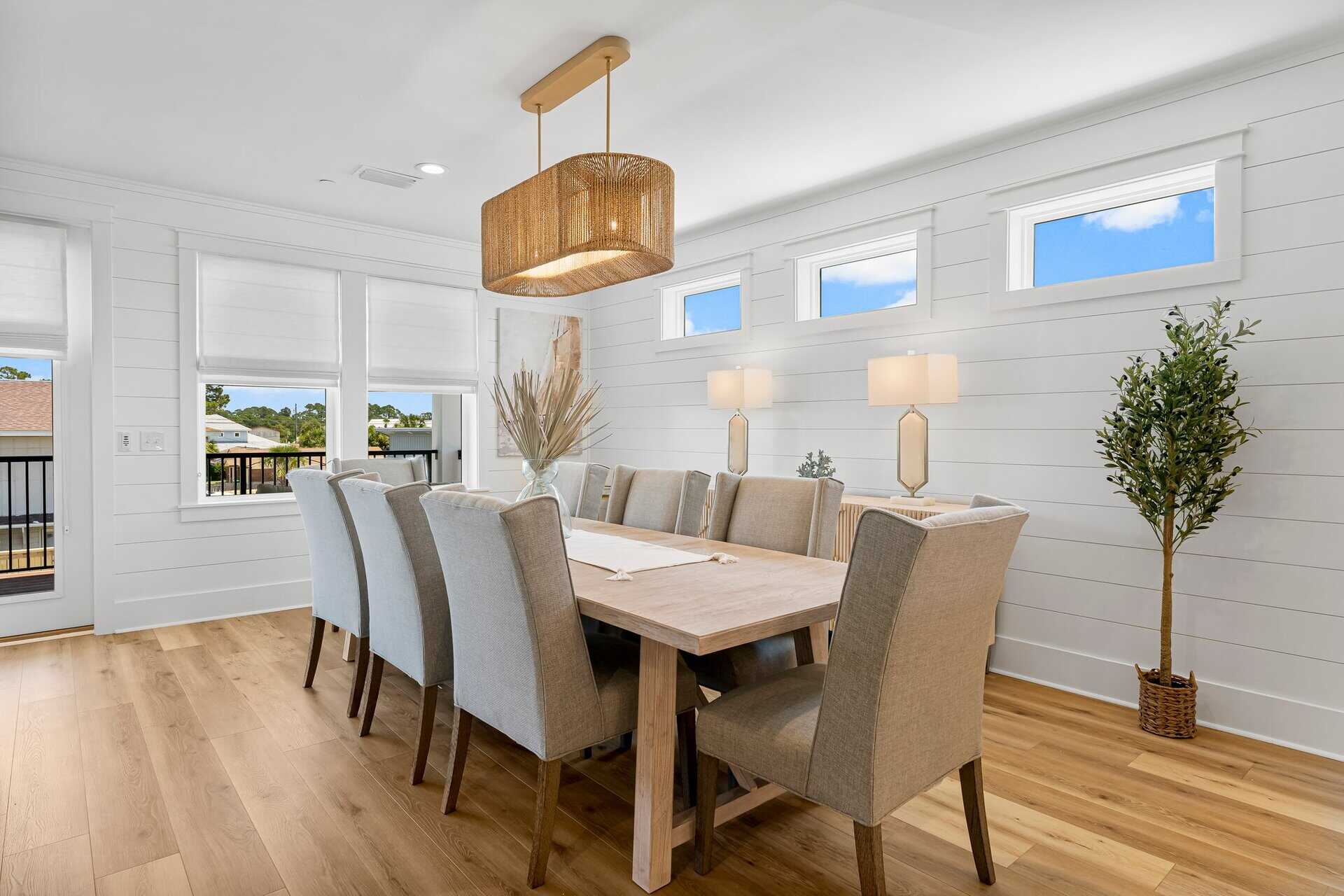 Bright dining room with wooden table, cushioned chairs, and modern pendant light. Large windows offer outdoor view; potted plants add greenery.