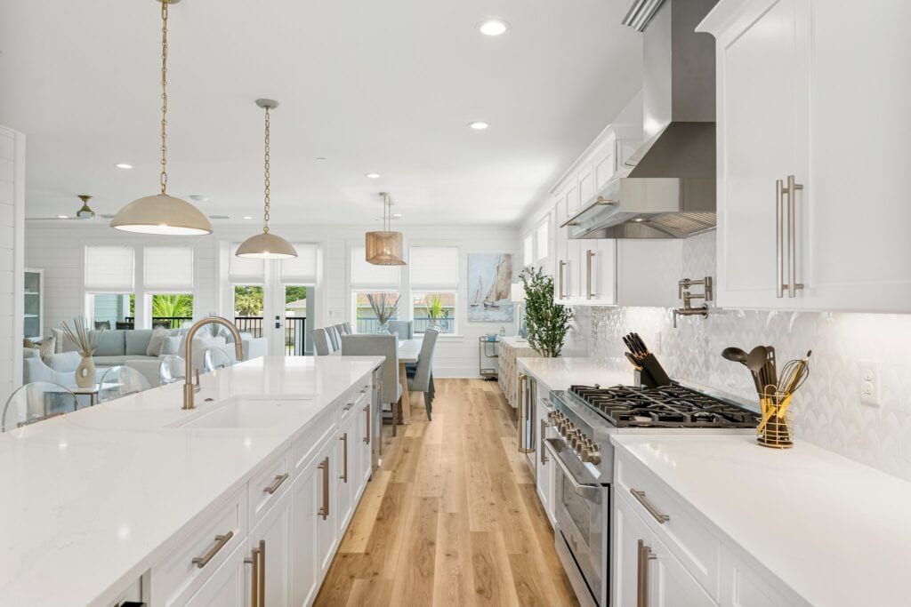 Spacious modern kitchen with white cabinets, large island, stainless steel appliances, and wooden flooring, leading to a bright dining and living area.