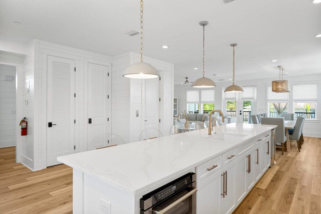 Spacious, modern kitchen features white cabinets, island with pendant lighting, wood flooring, adjacent dining area, large windows, and open living space. No visible people.