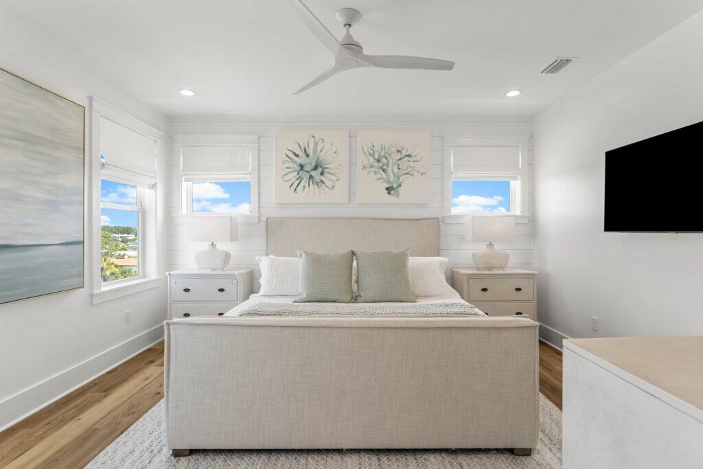 Bright, modern bedroom with neutral tones, stylish artwork above bed, wall-mounted TV, and scenic view through windows, featuring wooden floor and ceiling fan.