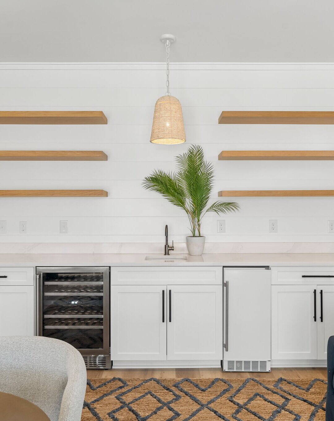 Modern kitchen with white cabinets, open wooden shelves, a potted plant, and pendant lighting. Includes a wine cooler and patterned rug.