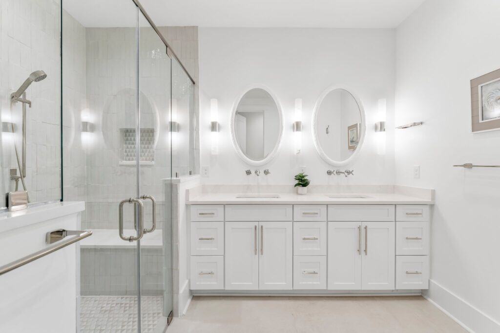 Modern bathroom with glass shower enclosure, double vanity, oval mirrors, and sleek white cabinetry. Minimalist design with bright lighting and decorative elements.