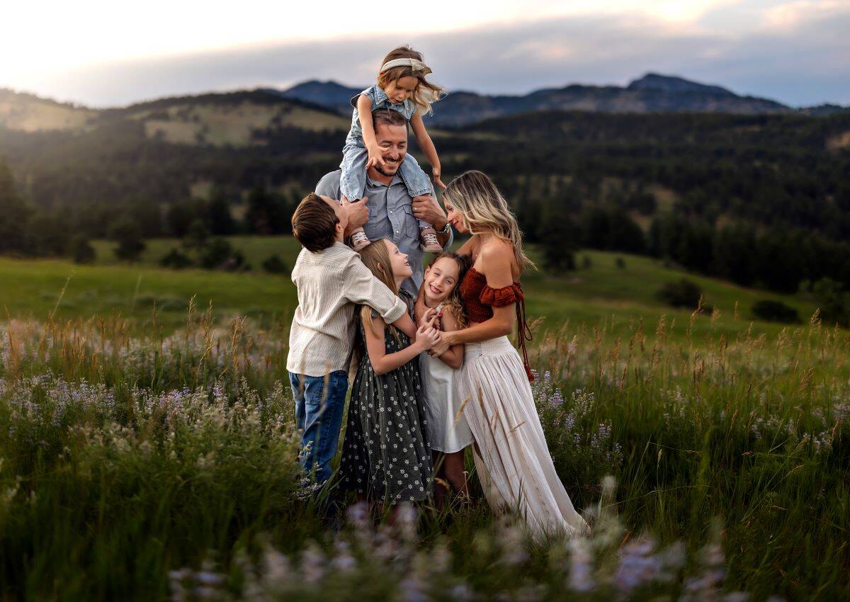 Five people joyfully embrace in a meadow with wildflowers, surrounded by rolling hills and distant mountains under a softly lit sky.