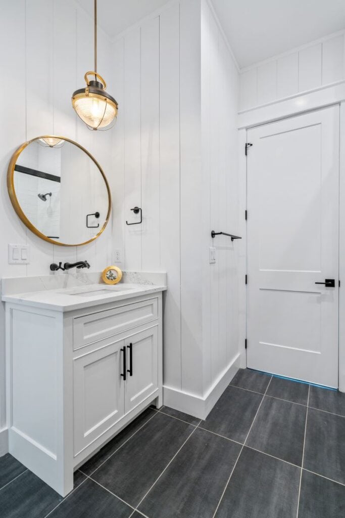 Minimalist bathroom with white panel walls, black fixtures, and modern lighting. Features a round mirror above a marble-topped vanity on dark tiled floor.