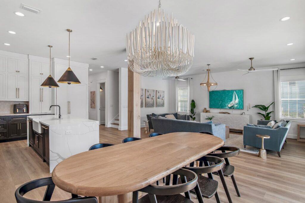 Modern open-concept living area with sleek kitchen, wooden dining table, and stylish lighting. Cozy seating facing a TV with ocean artwork.