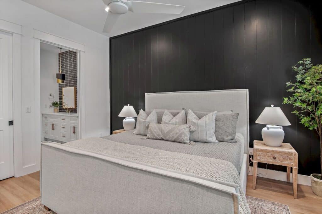 Modern bedroom with a large bed, black accent wall, ceiling fan, and adjacent bathroom featuring a vanity and stylish lighting.