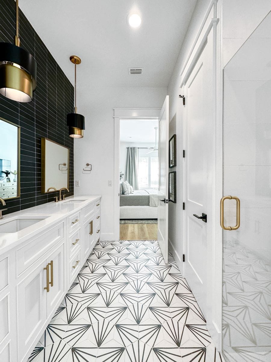 Modern bathroom with black and gold accents, geometric tile flooring, white cabinetry, and dual sinks, leading to a sunlit bedroom with gray decor.
