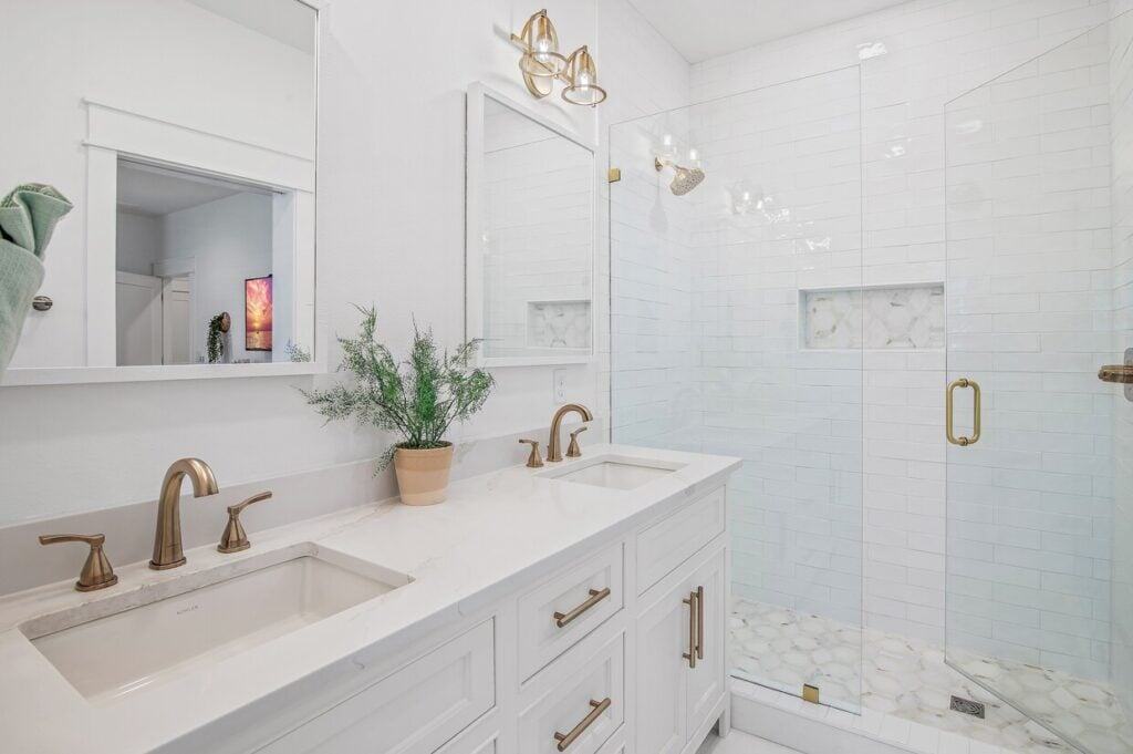 Modern bathroom with dual sinks, brass fixtures, glass shower, white tiles, wall mirrors, potted plant, folded towel, reflecting an inviting interior scene.