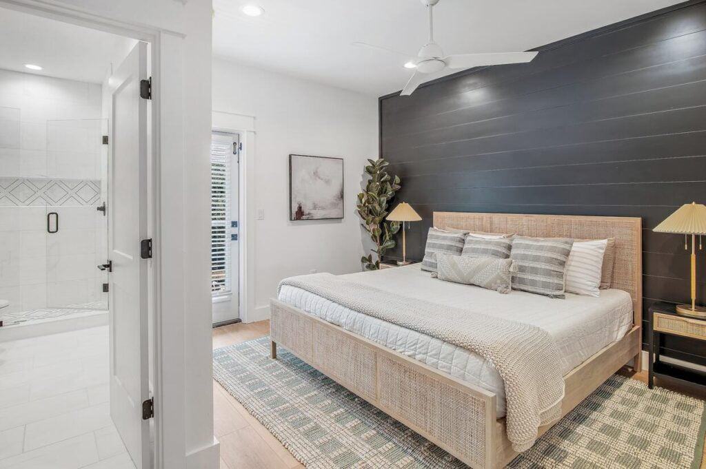 Modern bedroom with a large bed, dark accent wall, and ceiling fan. Bathroom visible through open door. Neutral colors and minimalist decor.