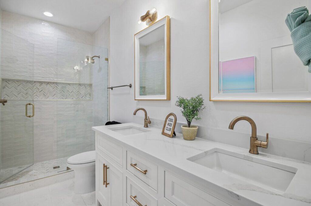 Modern bathroom with double sinks, gold fixtures, glass shower, potted plant, and a framed sign reading "Cool Kids Club" on the counter.