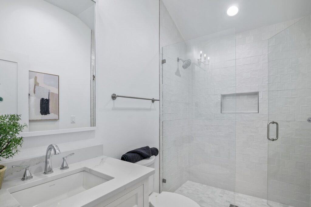 Modern bathroom with glass shower, white tiles, marble countertop, chrome fixtures, abstract painting, and decorative plant. Brightly lit, minimalistic design.