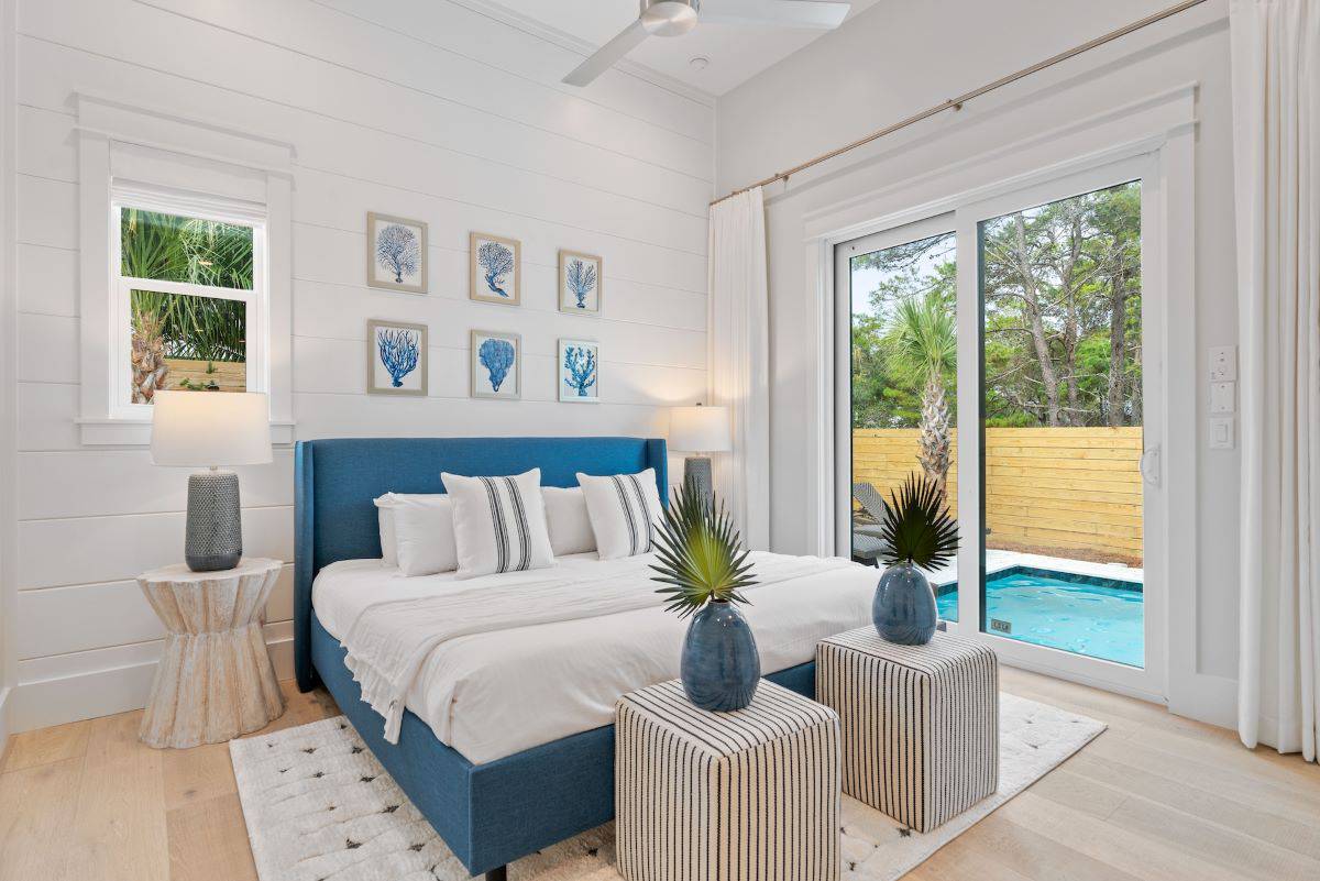 Bright, modern bedroom with a blue bed, white bedding, and sliding glass doors opening to a private pool. Minimalist decor with coastal artwork.